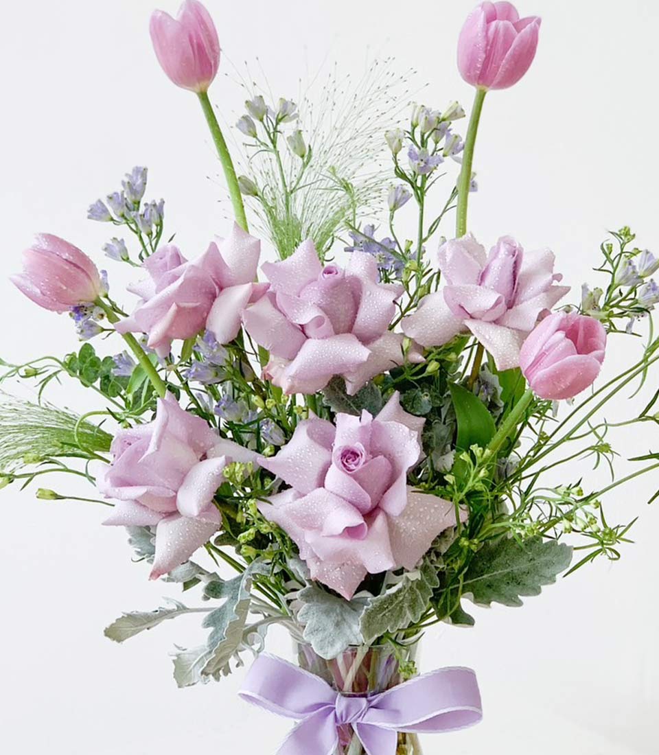 stunning flower bouquet in shades of purple, featuring roses and tulips in a glass vase, accented with lush greenery and tied with a purple ribbon