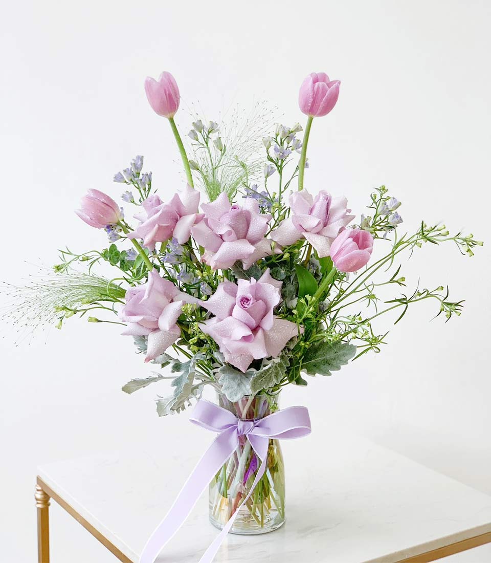 beautiful flower arrangement featuring light purple roses and pink tulips in a clear glass vase, adorned with a purple ribbon and lush greenery.