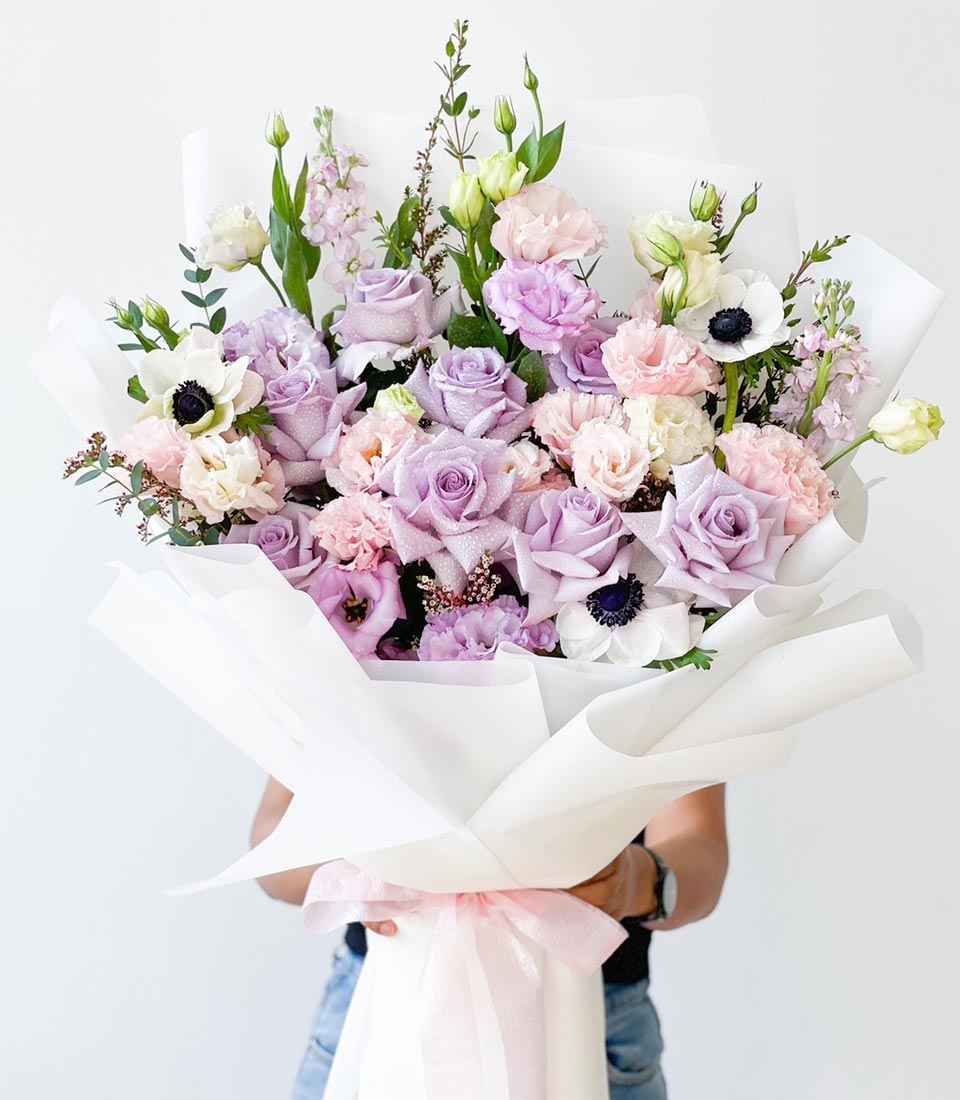 stunning bouquet of soft pink roses, light purple lisianthus, and white anemones, wrapped in white paper with a pastel pink ribbon, held by a person