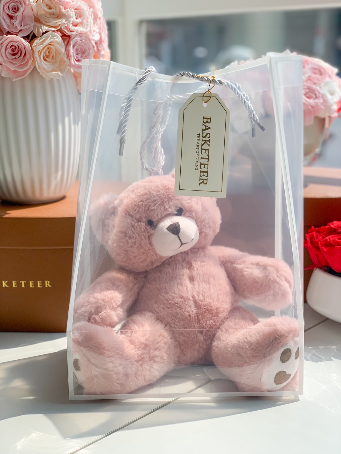 A soft blush pink teddy bear elegantly packaged in a transparent gift bag with a Basketeer tag. A perfect gift for babies, lovers, and special moments.