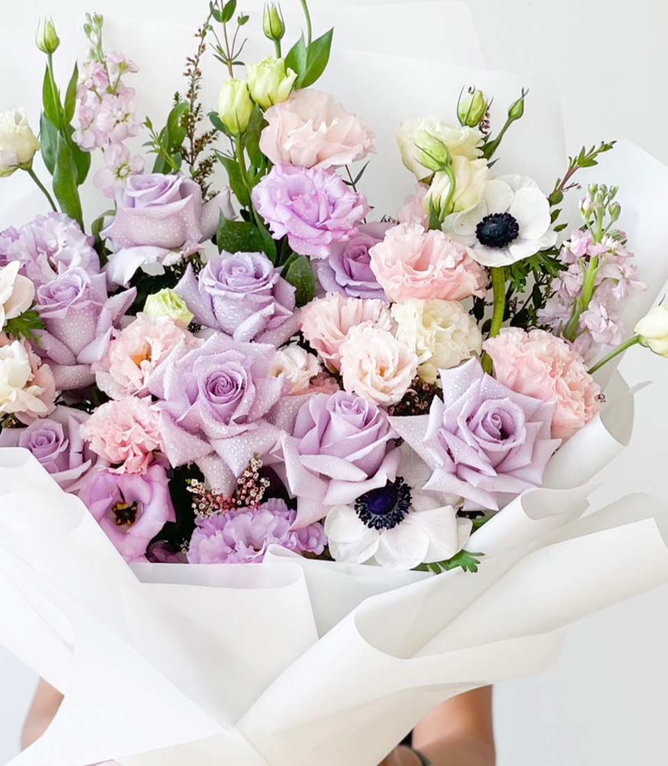 Elegant bouquet of pastel-colored flowers including roses, tulips, and greenery, beautifully arranged in a clear glass vase tied with a lavender ribbon.