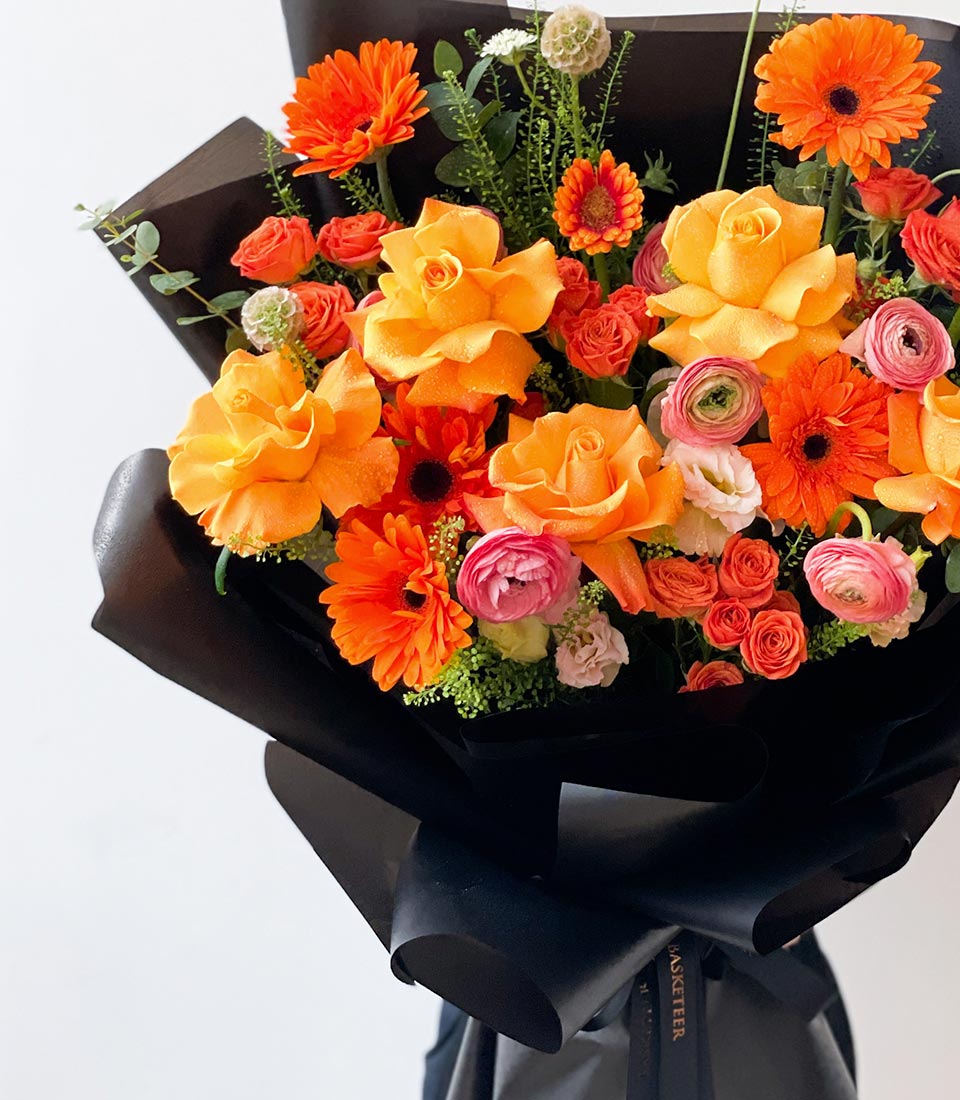 vibrant bouquet featuring a mix of orange roses, pink ranunculus, gerbera daisies, and greenery, wrapped in elegant black paper with a matching black ribbon.