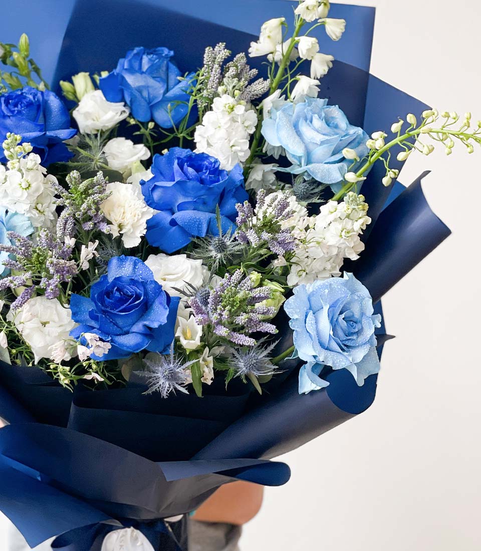 bouquet featuring blue roses, white carnations, lavender sprigs, and greenery, wrapped in blue paper.