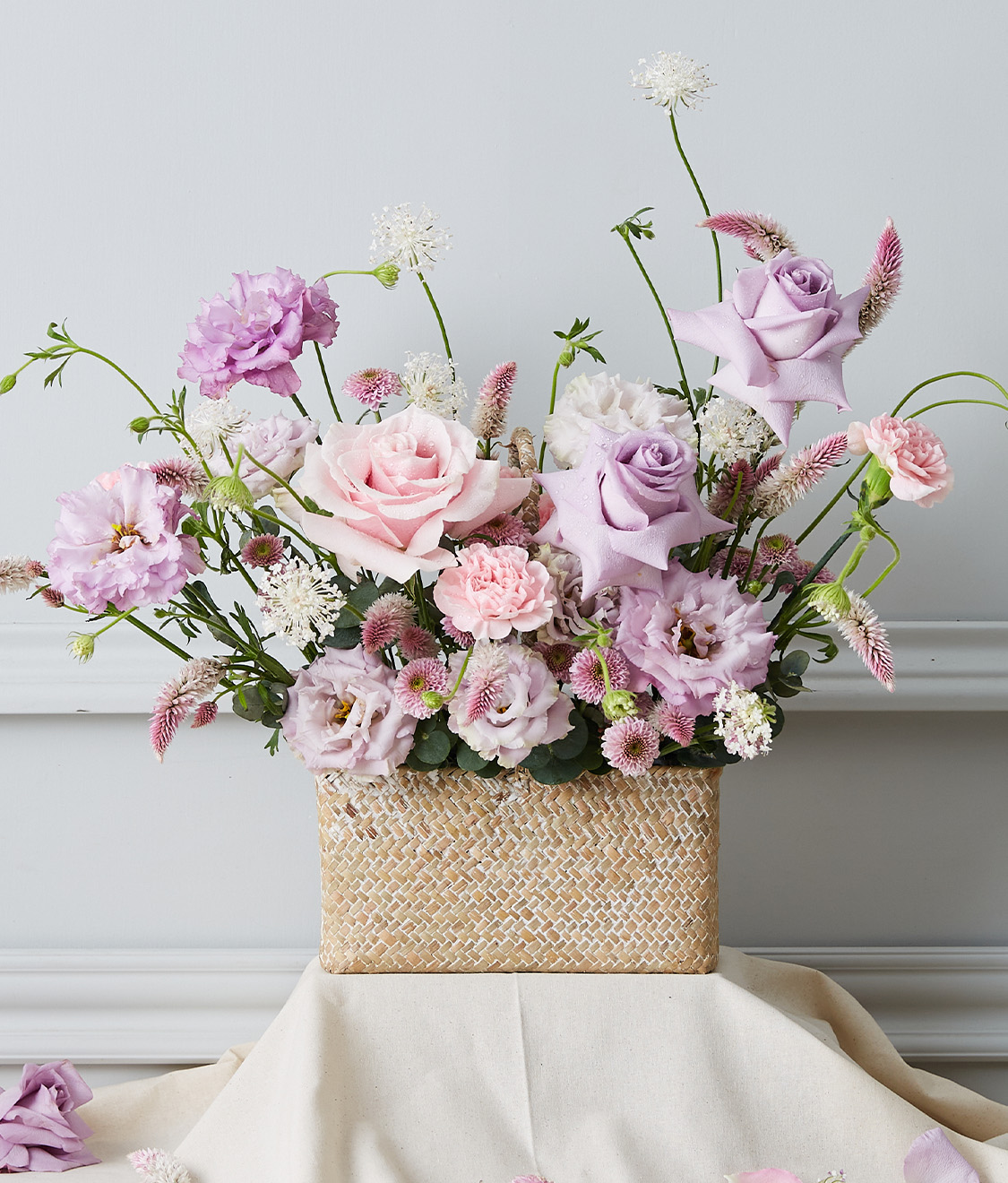 A sophisticated floral arrangement boasting an exquisite selection of pastel-hued blooms, featuring luxurious pink and lavender roses intertwined with delicate light purple flowers. This elegant bouquet is artfully presented in a finely woven basket, marked by the Basketeer logo on a chic ribbon. The scene is staged against an understated light grey wall, with scattered petals adding a touch of whimsical charm to the tableau.