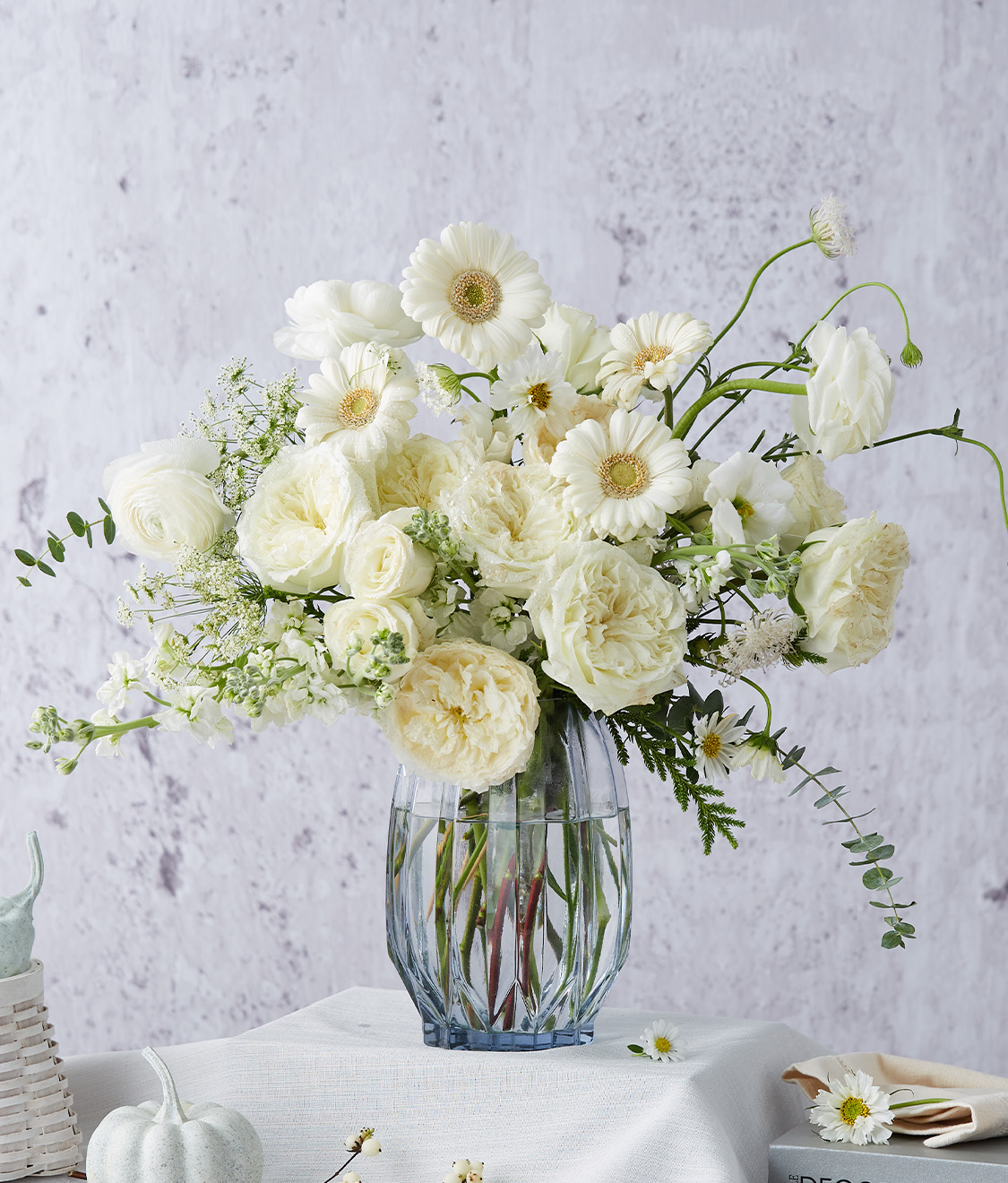 A glass vase cradles an exquisite arrangement of pristine white blooms, featuring elegant gerbera daisies and timeless roses, masterfully curated by Basketeer. The opulent display is set against a pure white tablecloth adorned with a tasteful stack of books and a chic green pumpkin, exuding sophistication. The setting is complemented by a textured light gray backdrop that enhances the luxurious ambiance. A discreet yet distinguished Basketeer ribbon graces the scene, symbolizing unparalleled quality and refinement.
