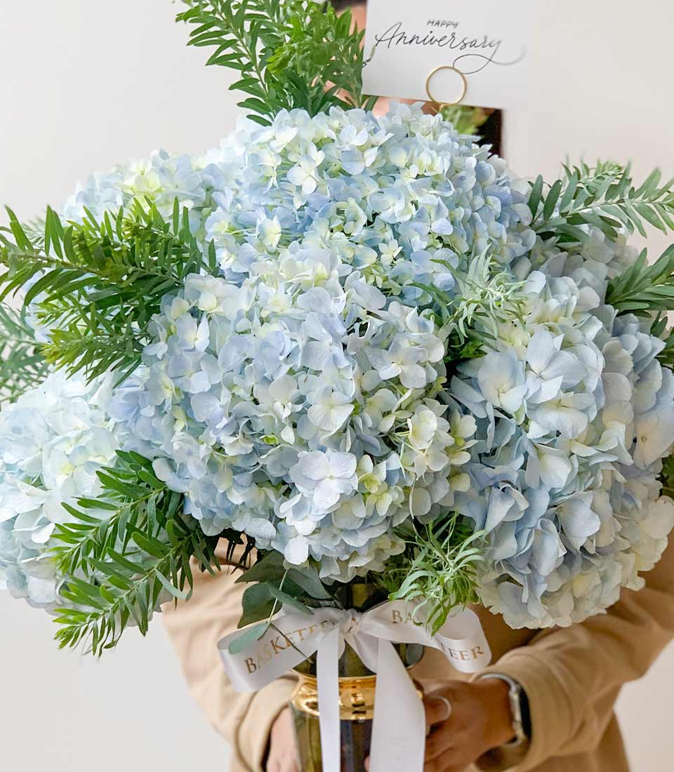 vibrant floral bouquet featuring blue hydrangeas and lush green foliage in a gold vase, celebrating an anniversary