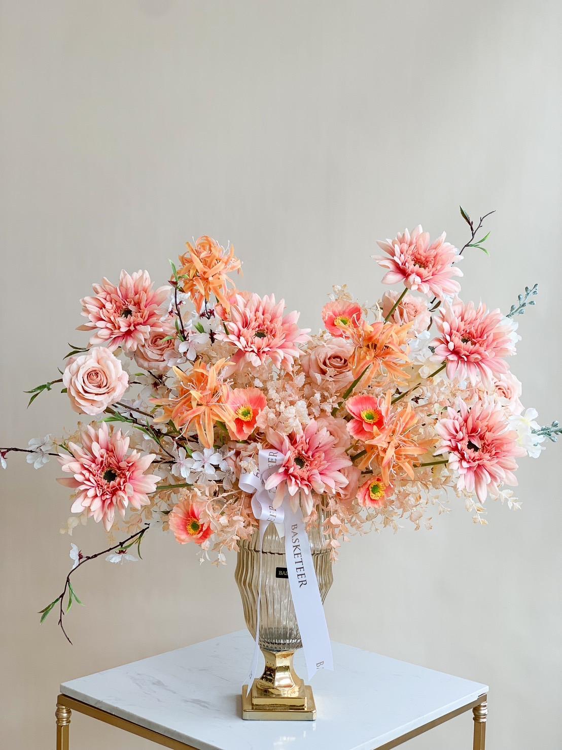 A large floral arrangement in a golden vase reminiscent of the Whispering Woods of Fall sits on a white marble table. The bouquet, consisting of pink and peach flowers like dahlias and roses, features white accents. A white ribbon with text is tied around the stems, all set against a plain beige background.