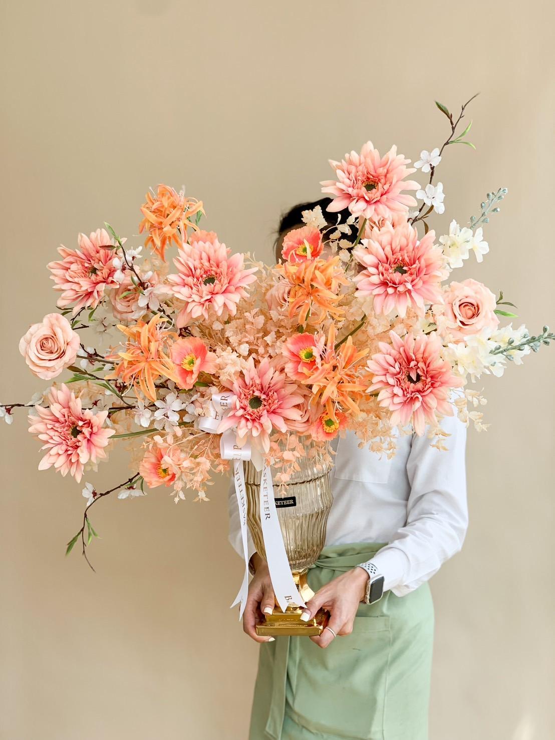 A person in a light shirt and green apron holds a tall, ornate floral arrangement, reminiscent of the Whispering Woods of Fall Artificial Flower Vase, with pink and orange dahlias and roses set against a neutral background.
