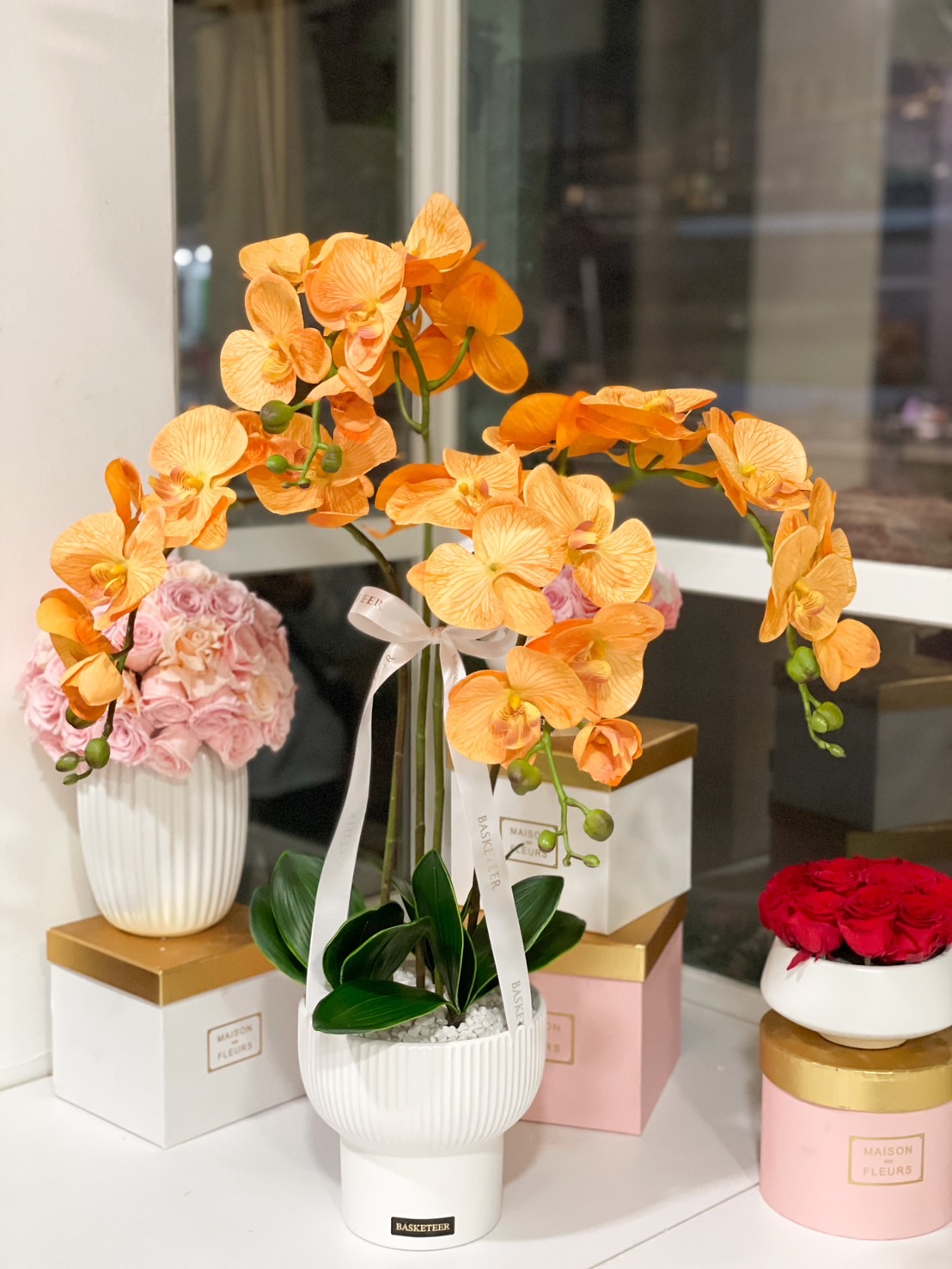 A vibrant golden-orange artificial orchid arrangement in a white ceramic vase, featuring realistic petals and lush green leaves.