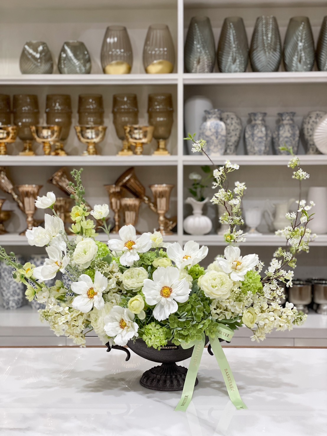 A stunning white floral arrangement featuring delicate white blooms, soft green hydrangeas, and blossoming branches in an elegant black urn vase. The perfect decorative piece for home, office, or special events.
