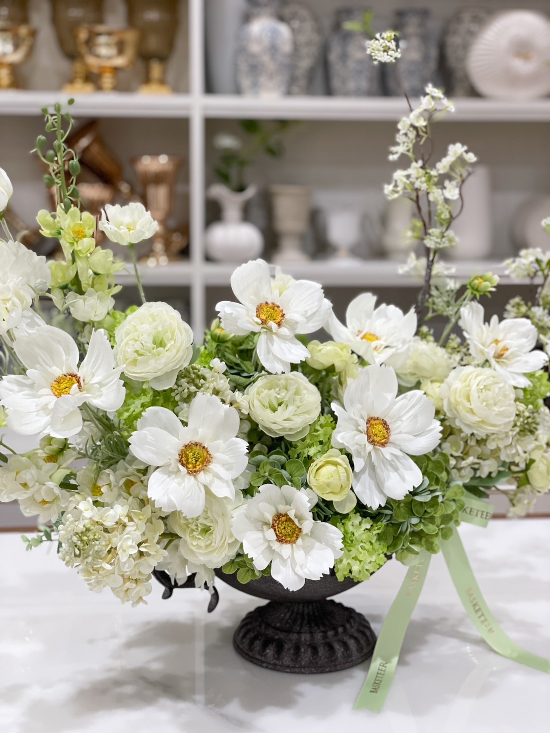 Close-up of a stunning white floral arrangement featuring delicate white blooms, soft green hydrangeas, and blossoming branches in an elegant black urn vase. The perfect decorative piece for home, office, or special events.