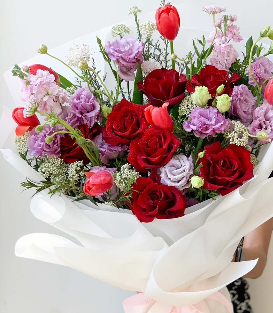 Elegant floral bouquet with red roses, purple lisianthus, and pink tulips wrapped in white paper, adorned with a pink ribbon.