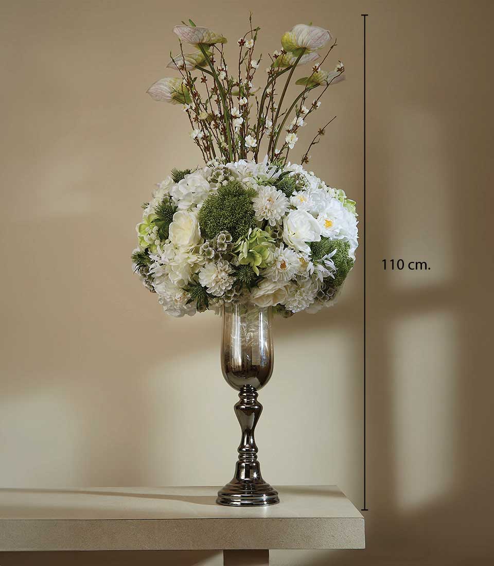 A luxurious floral arrangement by Basketeer graces a sophisticated setting, showcased in a tall, elegant dark glass vase. This opulent bouquet features an array of pristine white flowers interwoven with lush green accents and stately, budding branches, reaching an impressive height of 110 cm. The ensemble stands proudly against a beige wall bathed in soft, inviting lighting. A ribbon adorned with the Basketeer logo subtly enhances the scene's refined ambiance.