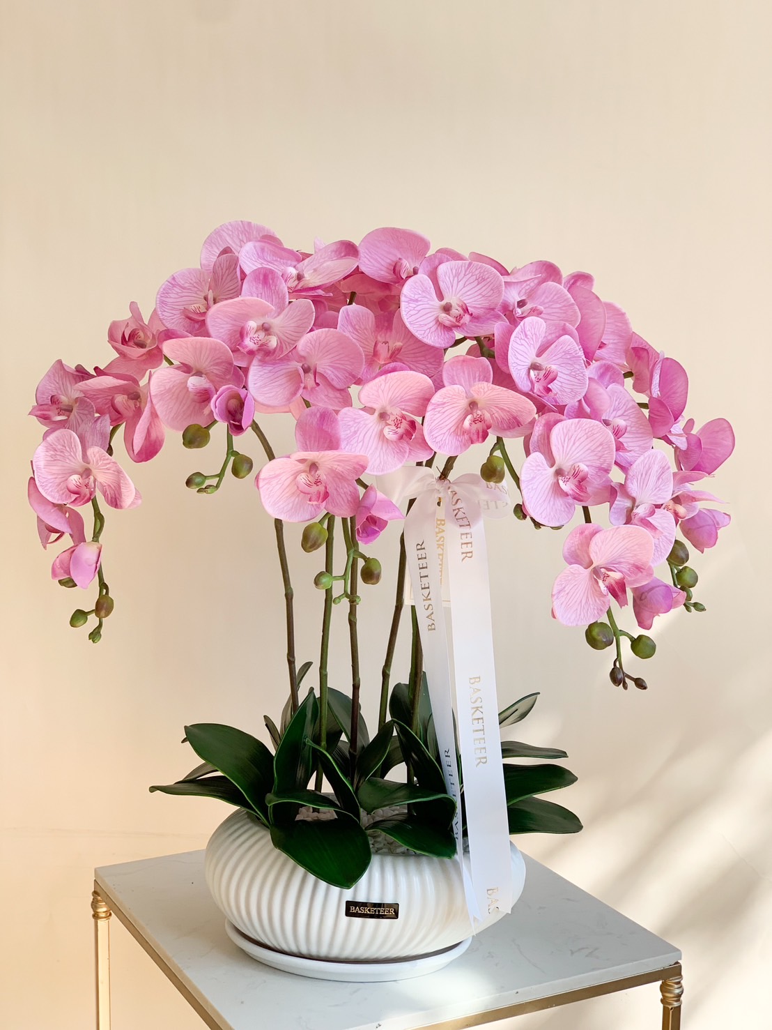 A gorgeous arrangement of pink Phalaenopsis orchids in a white ceramic vase with lush green leaves. A perfect decor piece for offices, homes, and events.