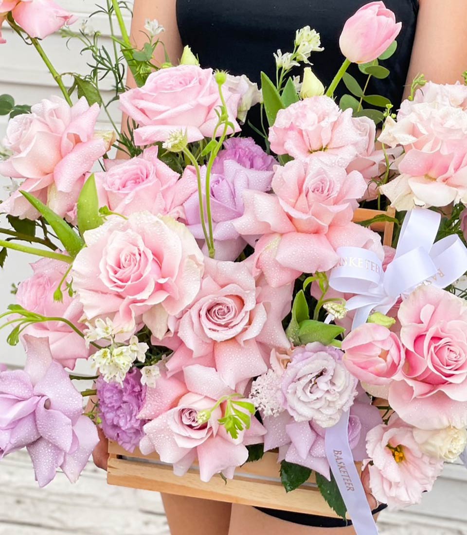 Beautiful arrangement of pink and lavender roses in a wooden crate, held by a person in a black dress, showcasing the elegance and charm of the delicate flowers. Perfect for special occasions and thoughtful gifts.