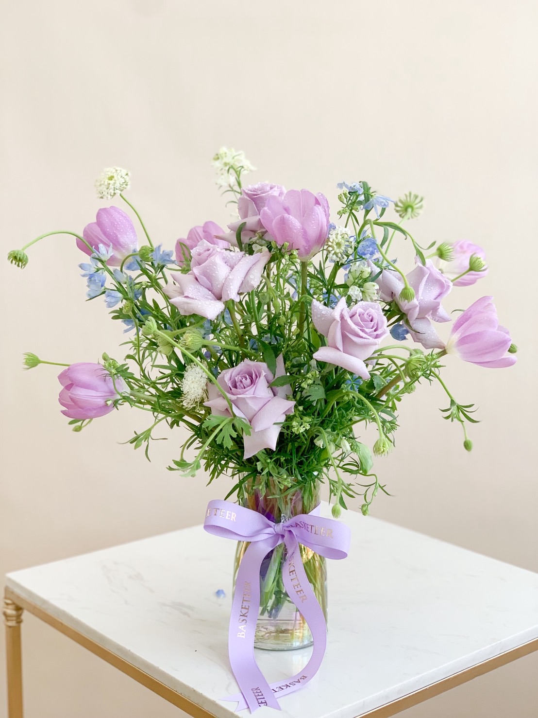 Vase arrangement of pastel purple roses and tulips with soft greenery and lavender ribbon.