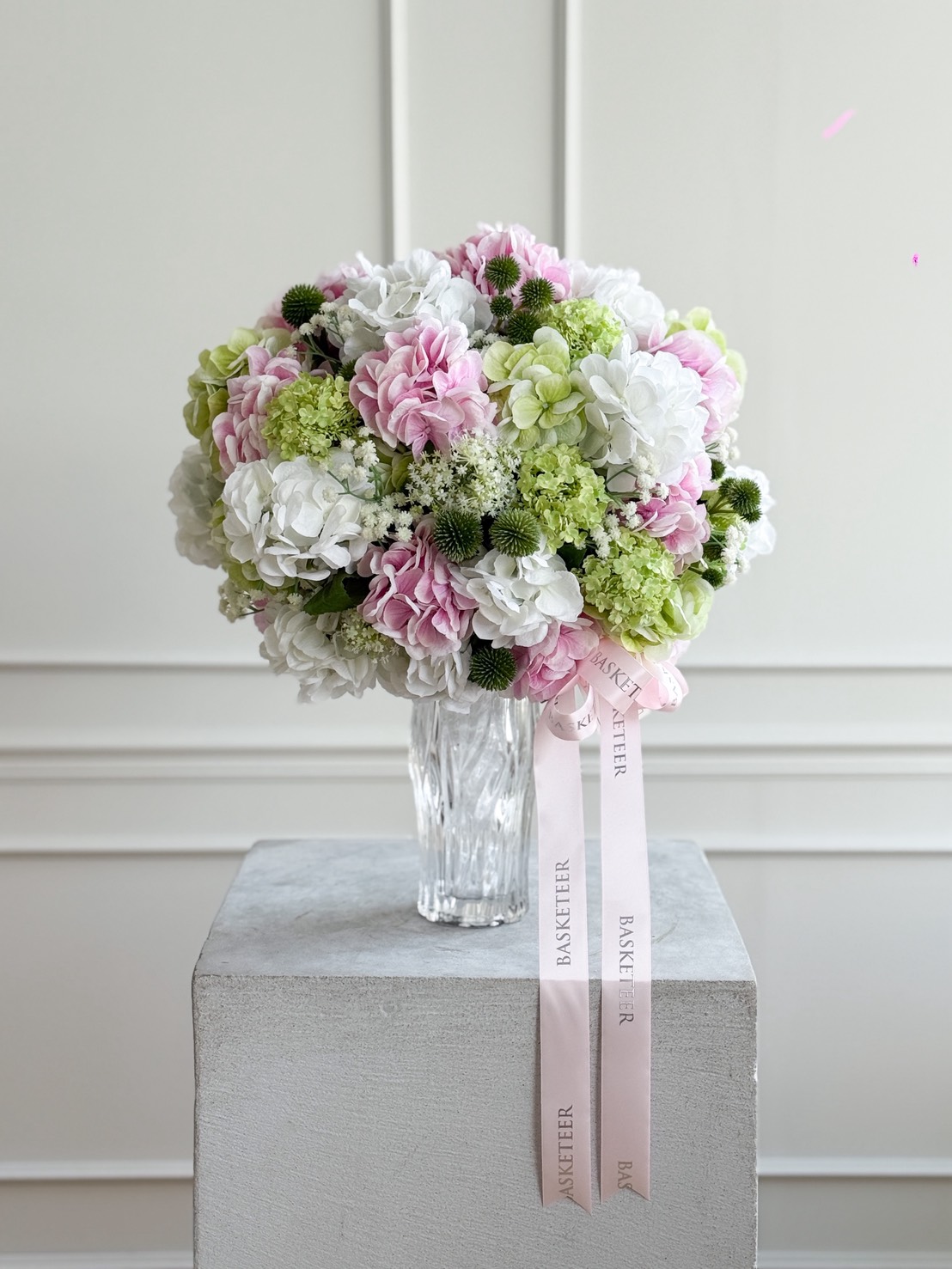 Pastel hydrangea flower arrangement in a crystal glass vase with pink and white blooms, green accents, and pink Basketeer ribbon.
