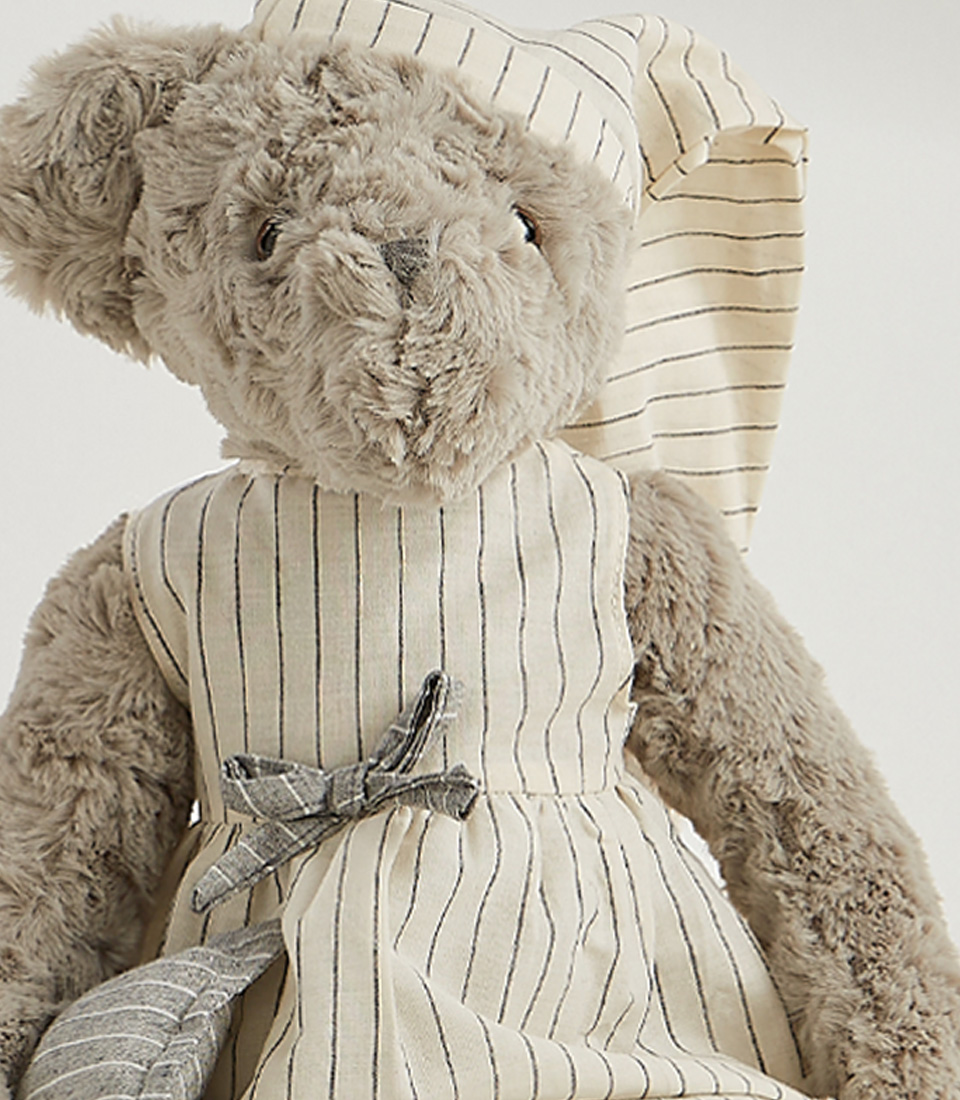 A grey teddy bear wearing a striped nightgown and matching nightcap, standing and ready for bedtime, set against a light gray background.