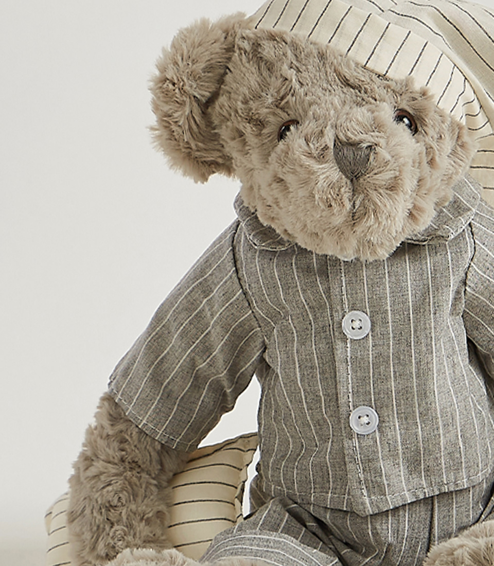 A grey teddy bear wearing striped pajamas and a matching nightcap, standing and ready for bedtime, set against a light gray background.
