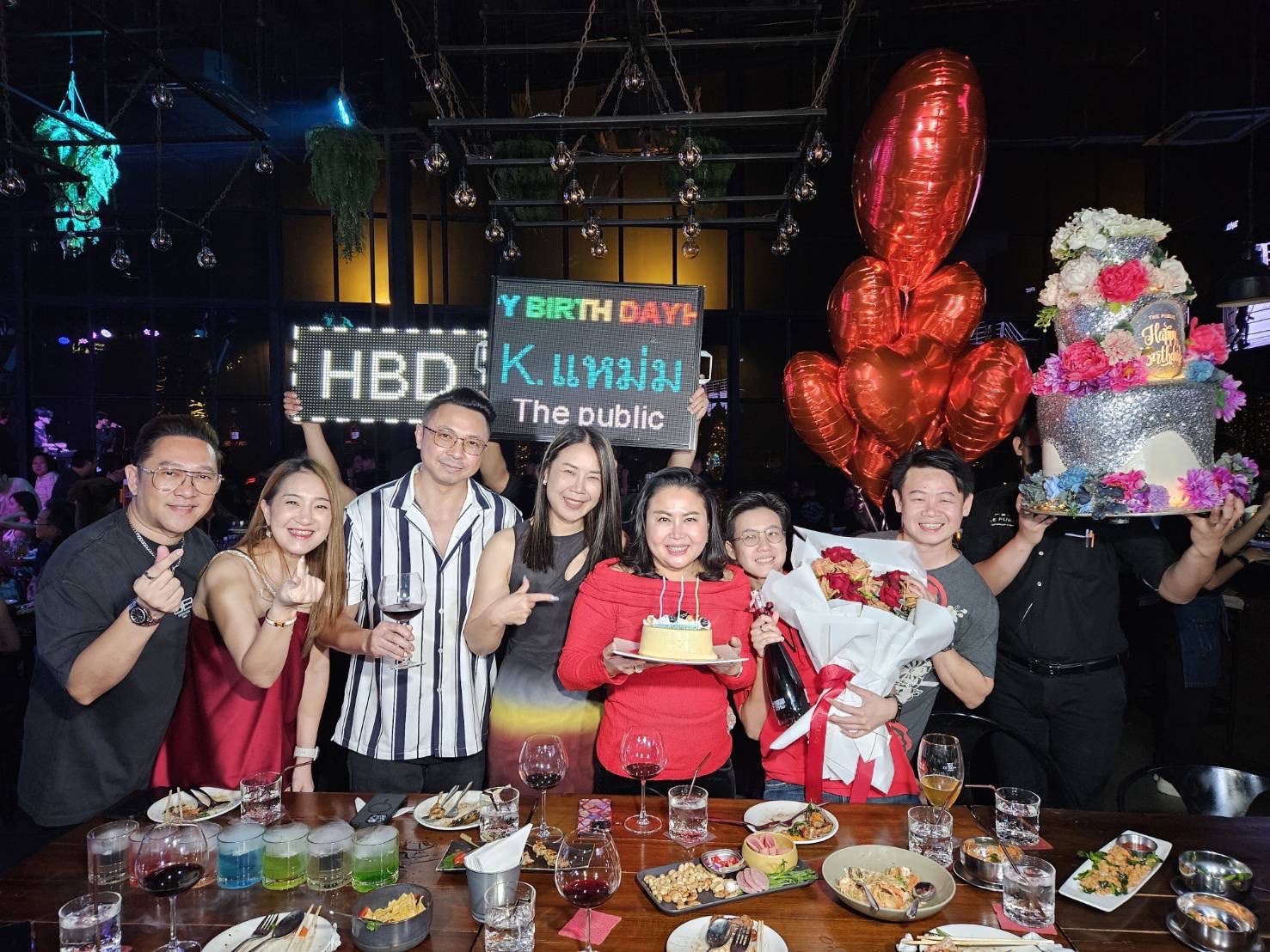 A group of people celebrate a birthday at a restaurant. They are posing for a photo behind a table with food and drinks. One person holds a cake, another has a bouquet of flowers, and two large balloon bouquets are visible. A sign reads 