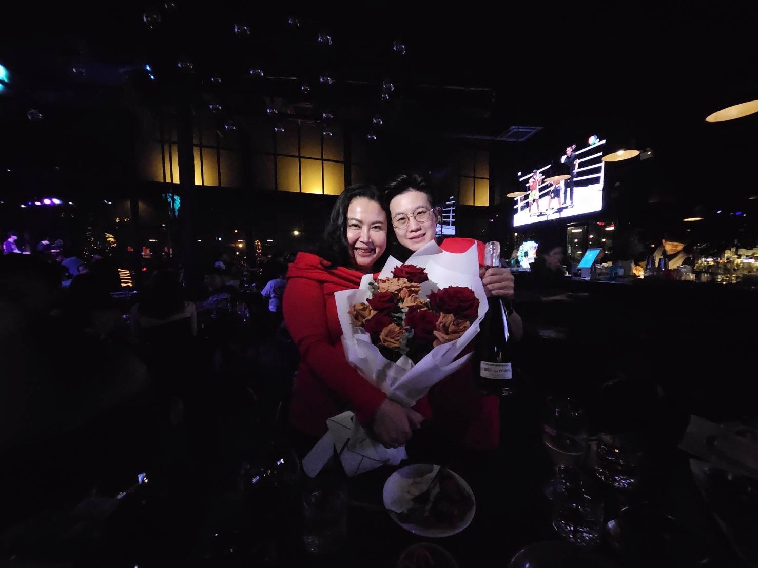 Two people are smiling and posing together in a dimly-lit venue. They are holding a large bouquet of red and white flowers and a bottle of wine. In the background, there are other people, illuminated tables, a bar, and a screen showing a sports match.