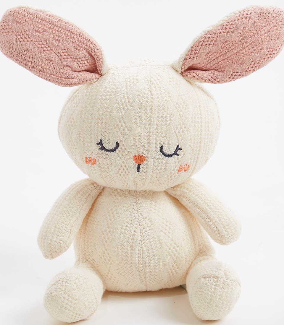 Close-up view of a handcrafted crocheted bunny plush toy, with a soft cream exterior and pastel pink ears. The bunny has a sweet, sleepy face with embroidered eyes and an orange nose, perfect for newborns and young children.