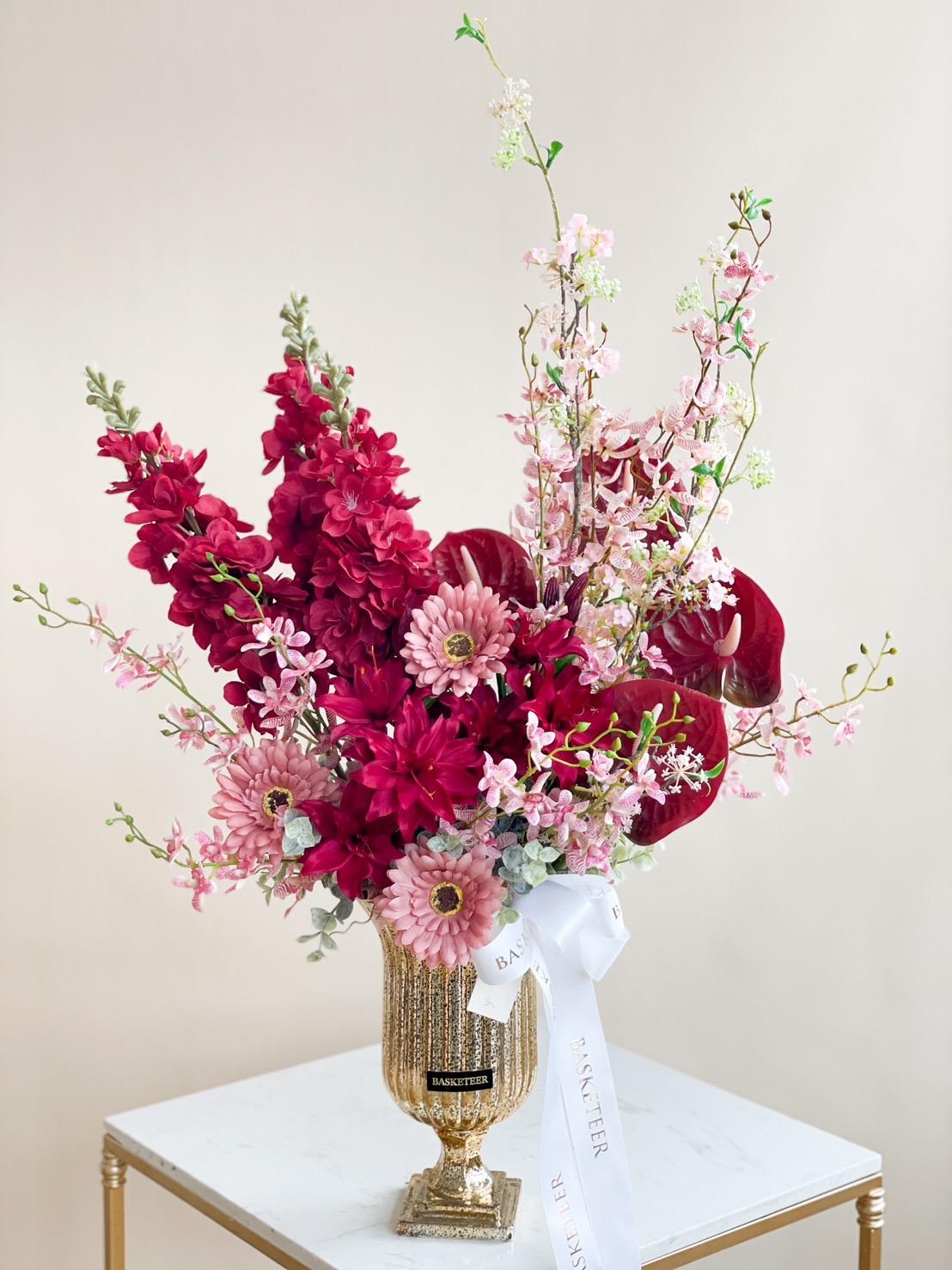A Scarlet Luxury gold vase sits on a white marble table, filled with an arrangement of pink and red flowers, including dahlias, snapdragons, and orchids. A white ribbon is wrapped around some stems, and the background is a neutral light beige color.