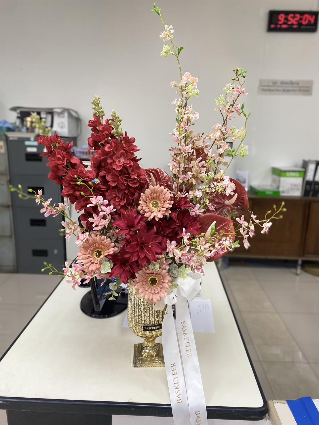         A decorative vase filled with a vibrant floral arrangement sits on a table. The bouquet features deep red, pink, and white flowers with lush greenery, complemented by a white ribbon. This Scarlet Luxury Artificial Flower Vase adds elegance to the office setting, where furniture and a digital clock show 9:52 AM.