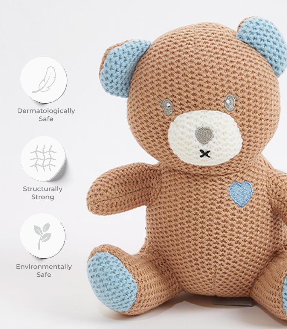 Close-up of an adorable brown crocheted teddy bear with blue accents on its ears, paws, and a heart detail on its chest. The bear is labeled as dermatologically safe, structurally strong, and environmentally safe.
