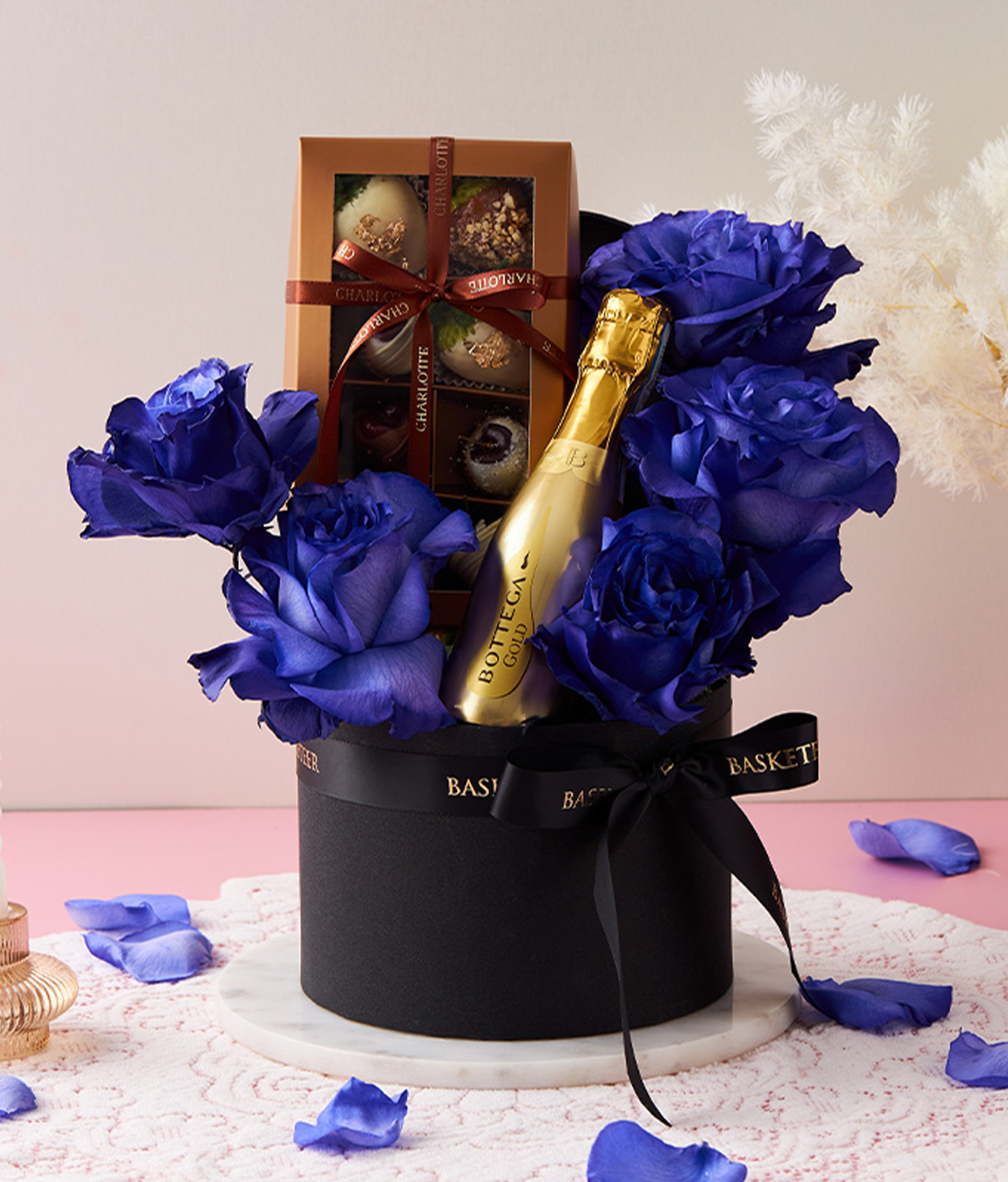 A black hatbox filled with five vibrant royal blue roses, a gold bottle of Bottega Prosecco, and a box of assorted chocolates tied with a brown ribbon. The arrangement is adorned with a black ribbon imprinted with 