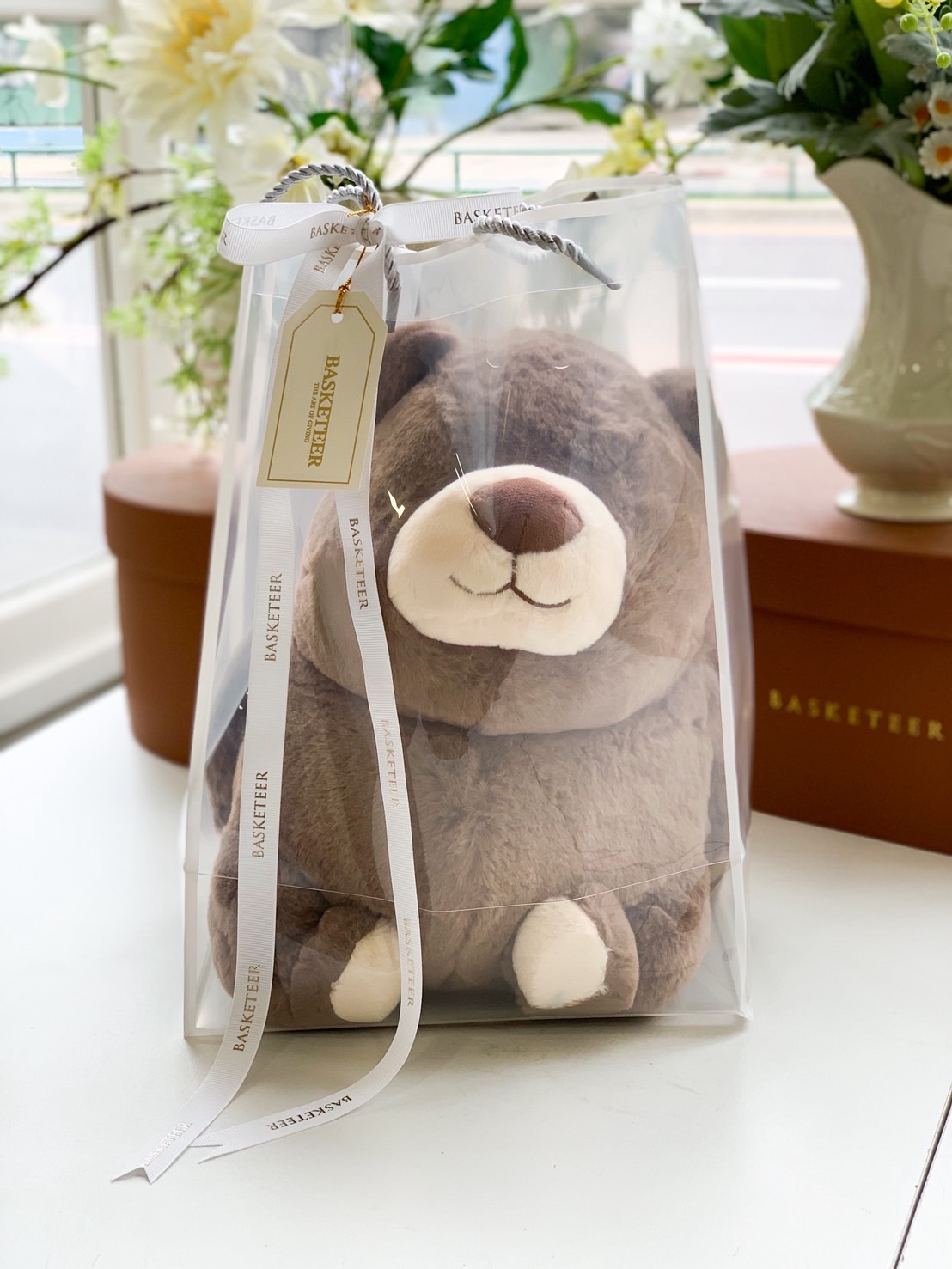 Plush brown teddy bear in a clear plastic gift box with white ribbon and gold-tagged label from BASKETEER