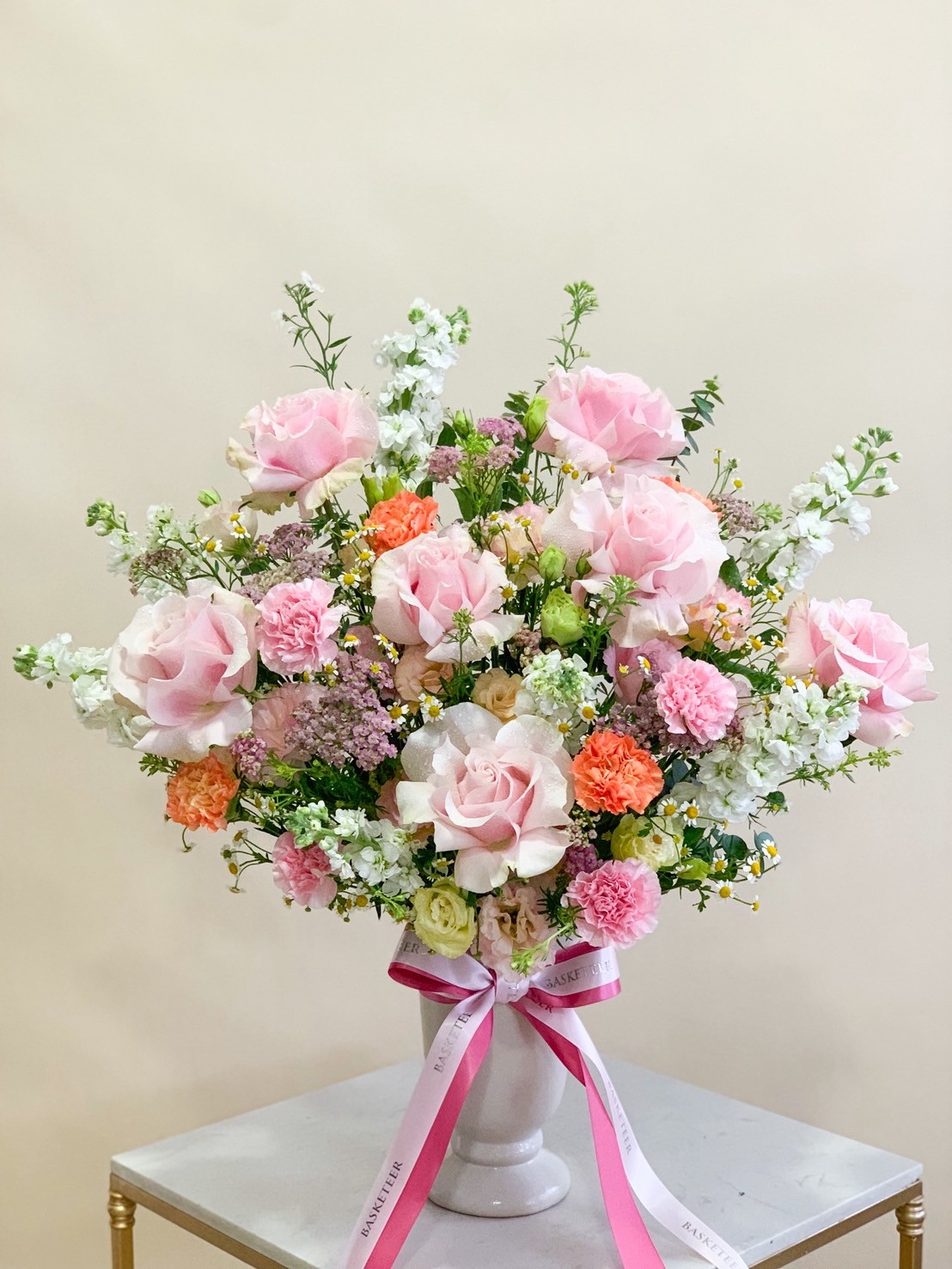Pedestal flower arrangement with large pink roses, carnations, white stock, orange blooms, and assorted pastel fillers tied with pink ribbons.
