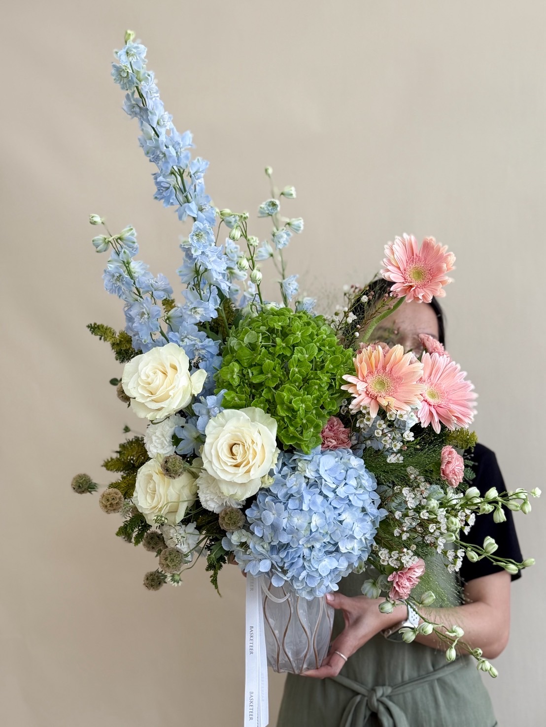Elegant floral vase with blue delphinium, green and blue hydrangeas, white roses, pink gerberas, and filler flowers in pastel tones