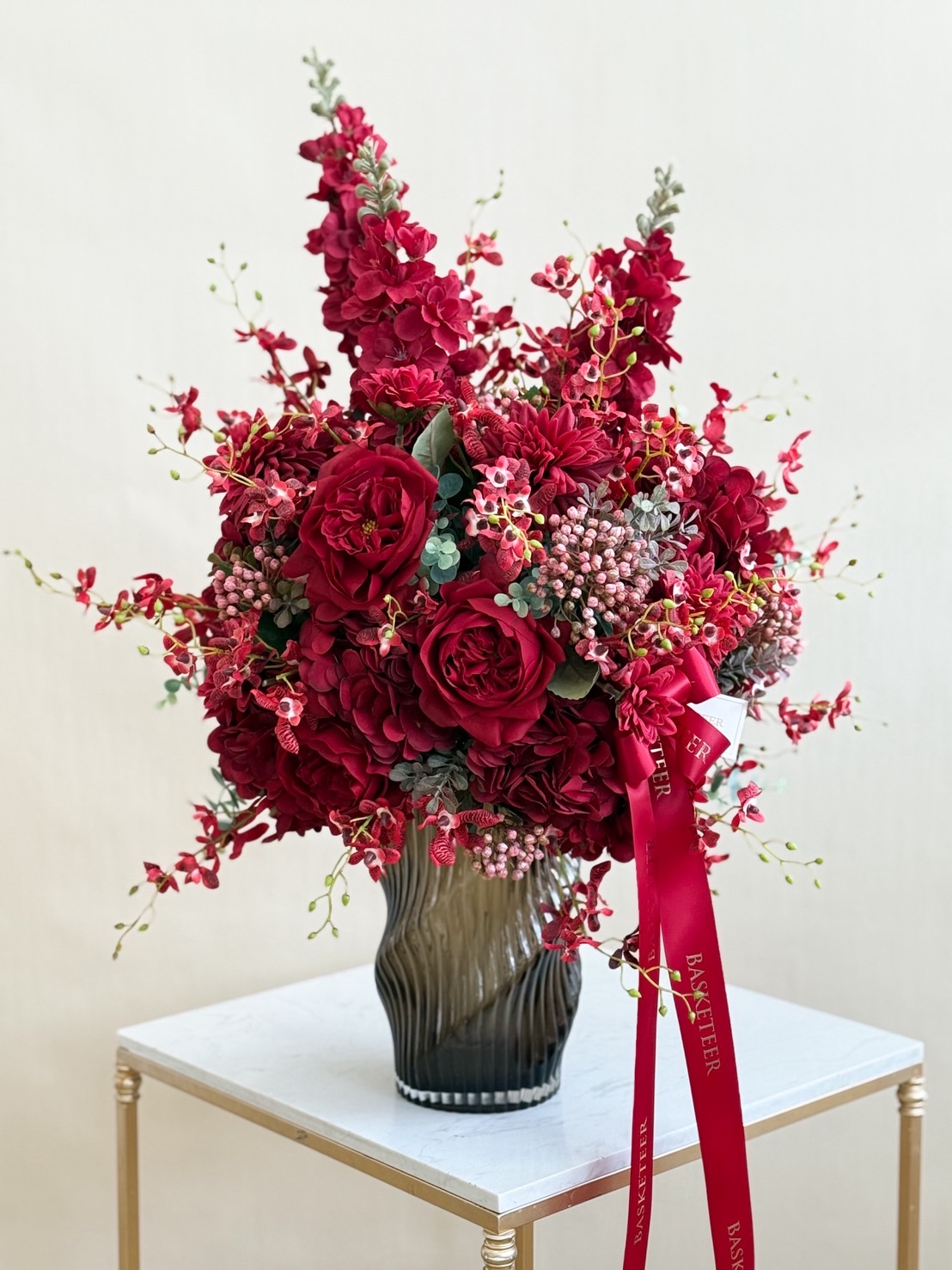 Elegant flower arrangement with red roses, orchids, and mixed red blooms in a tall fluted glass vase – luxury floral gift.