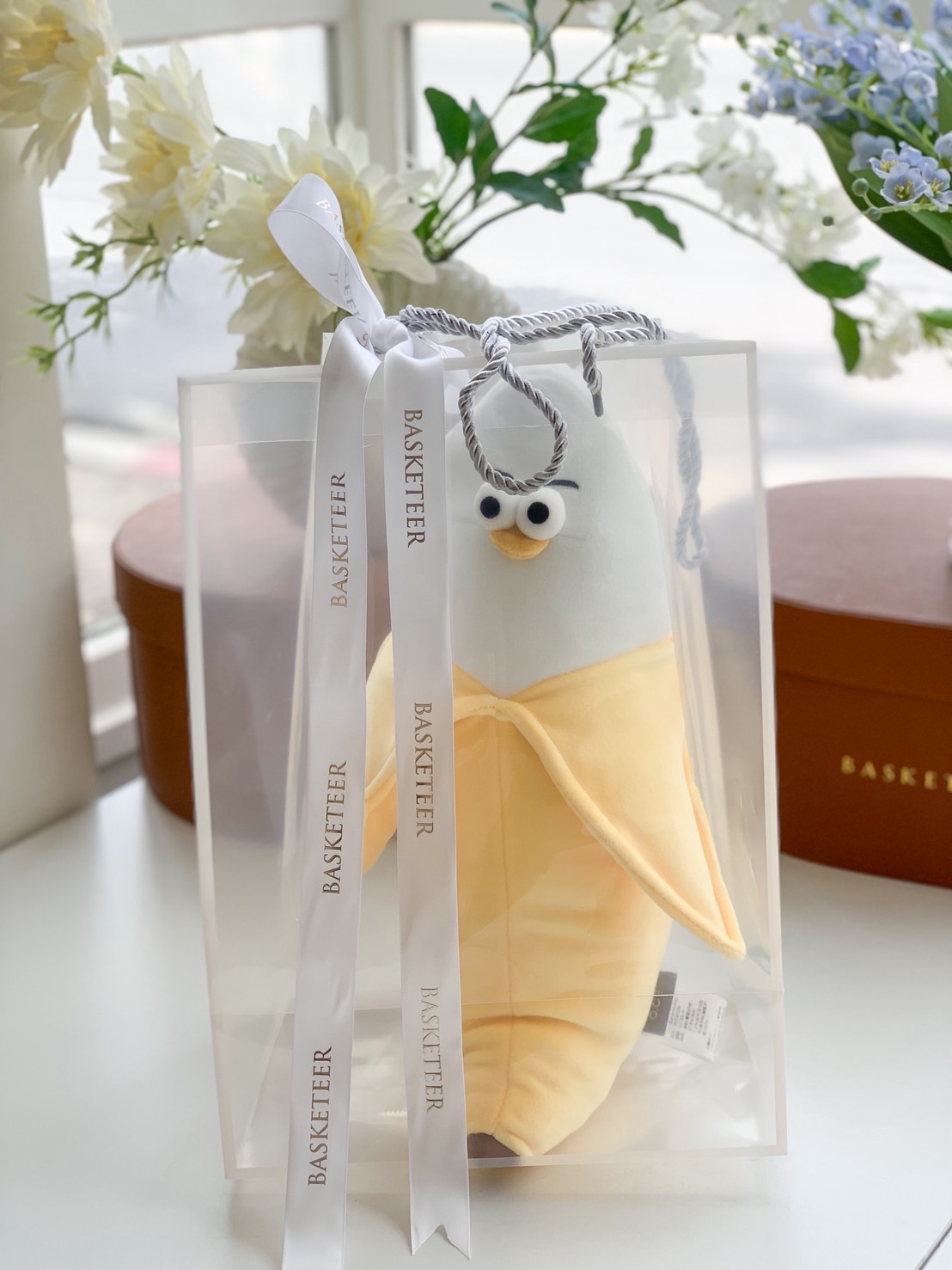 Cute penguin plush toy dressed in a banana peel suit, presented in a clear gift bag with white ribbon.