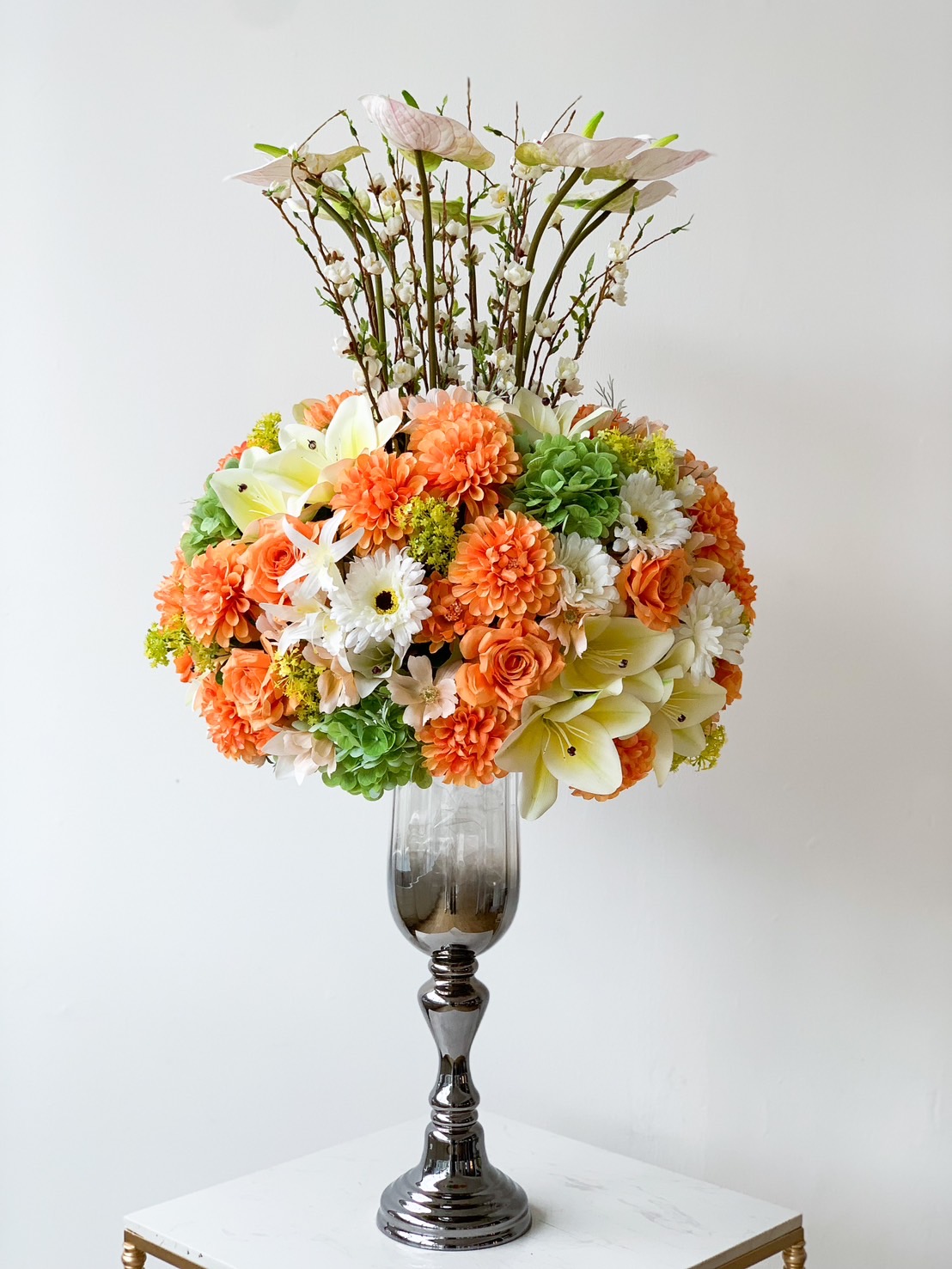 A tall, elegant glass vase holds a luxury floral arrangement with a variety of flowers, including orange carnations, white daisies, yellow lilies, and green hydrangeas, arranged in a round shape. Delicate branches extend upward from the center, adding height to the composition.