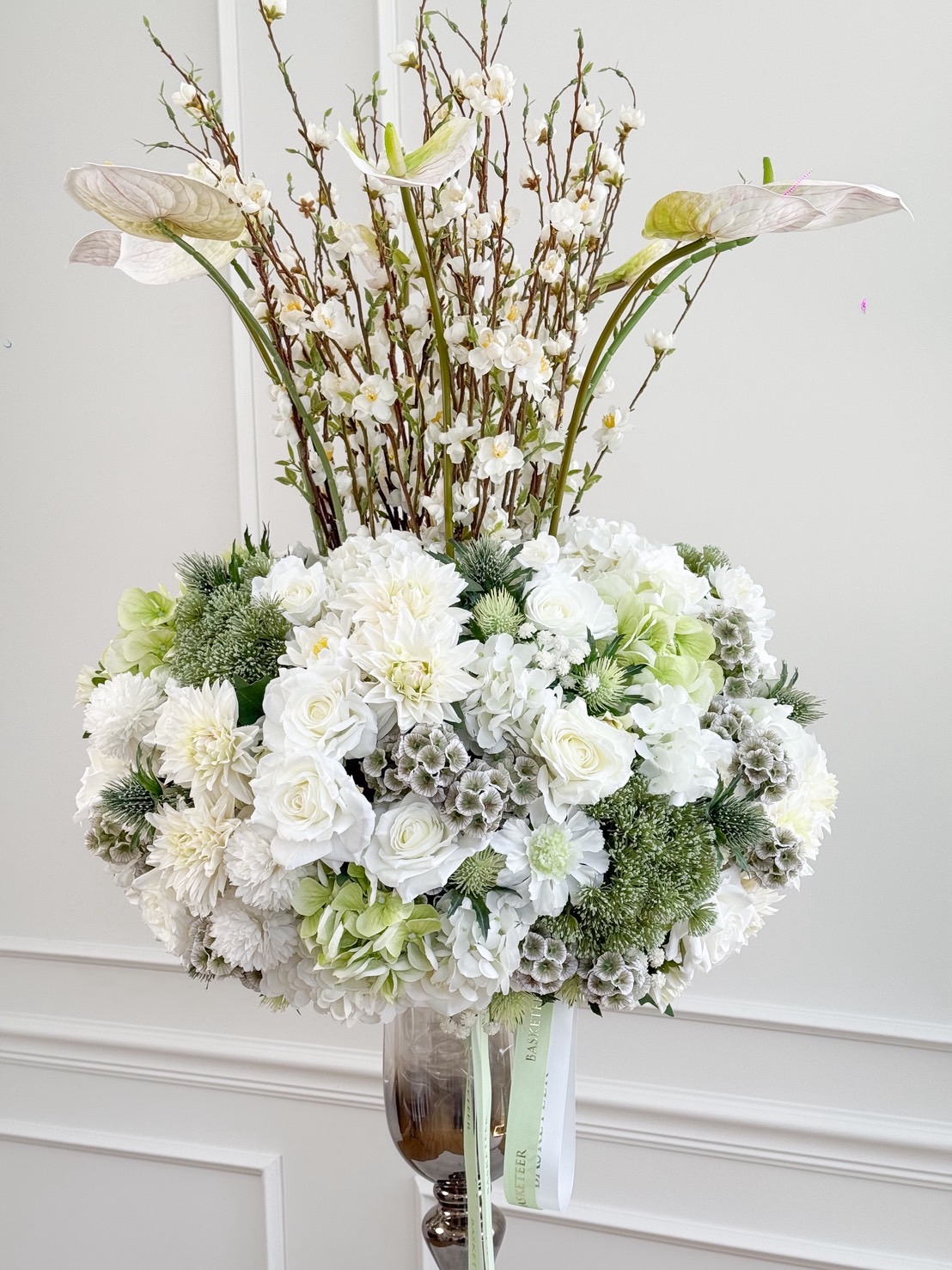 Luxury tall vase arrangement with white roses, anthuriums, hydrangeas, and flowering branches in silver pedestal glass.