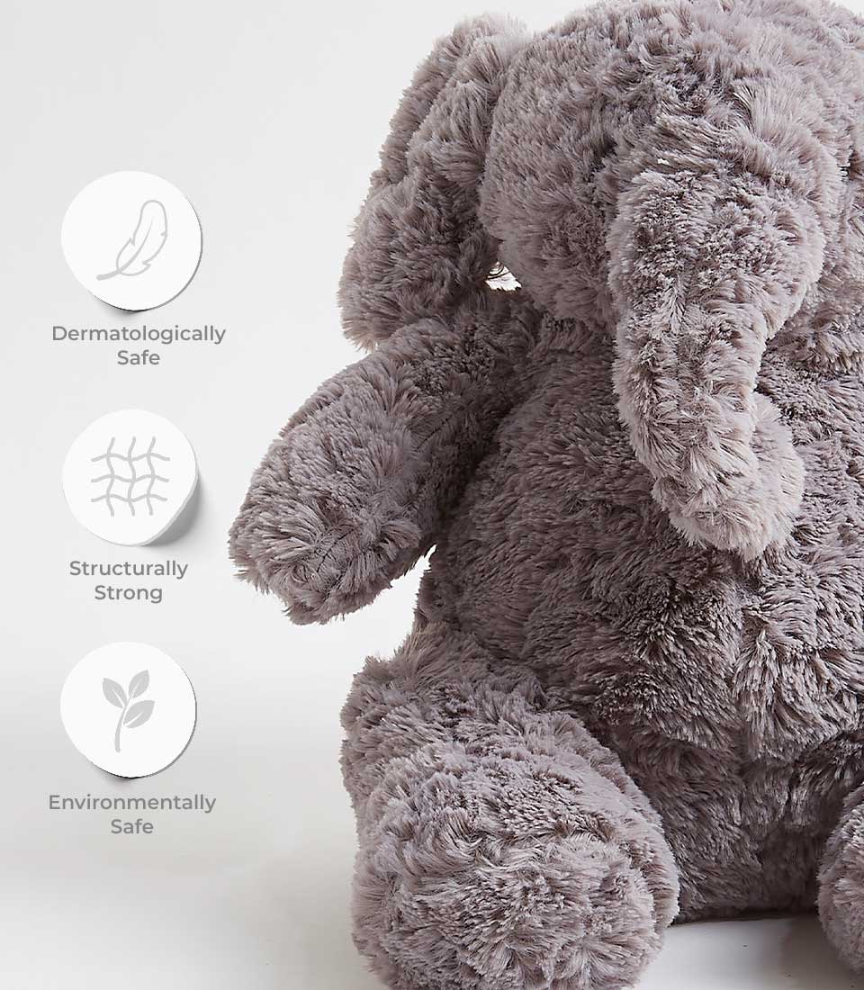 A fluffy, grey stuffed elephant toy is pictured on a white background. Three icons with text indicate the toy's features: 