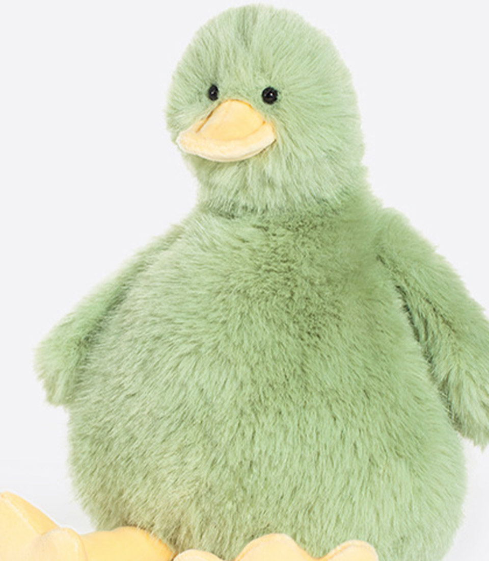 A round, plush green duck with a soft, fluffy texture is sitting upright. The duck has a yellow beak and black eyes. Its wings are short, and it has tiny yellow feet peeking out at the bottom. The background is a plain, light grey color.