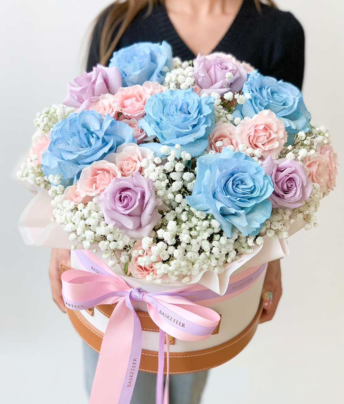 A person is holding a round hatbox filled with a bouquet of blue, pink, and lavender roses, accented by white baby's breath flowers. The hatbox is adorned with a pink ribbon bow and is labeled with the text 