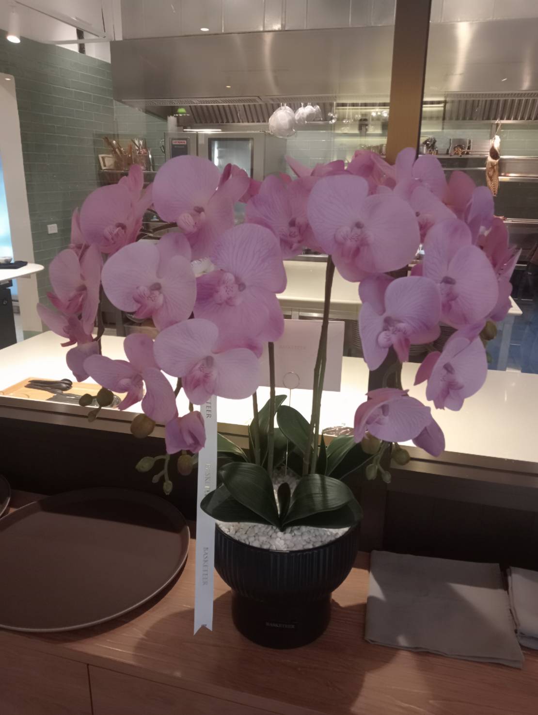 An elegant arrangement of light purple artificial orchids with broad petals is displayed in a black pot on a counter in a modern kitchen with stainless steel surfaces and various kitchen items in the background.
