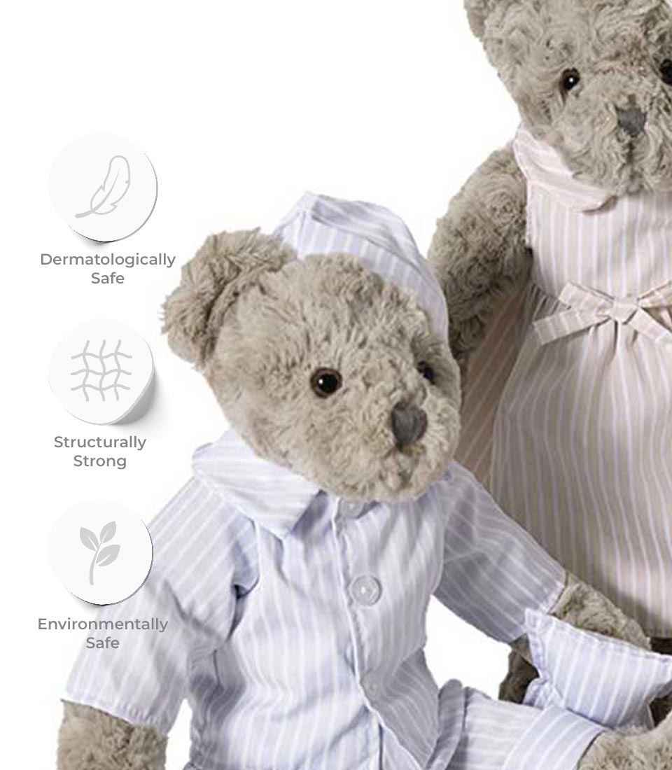 Two plush toy bears are shown dressed in striped clothing. The smaller grey teddy bear wears striped pajamas, while the larger bear dons a striped nightgown. Icons to the left indicate these soft toys are dermatologically safe, structurally strong, and environmentally safe.