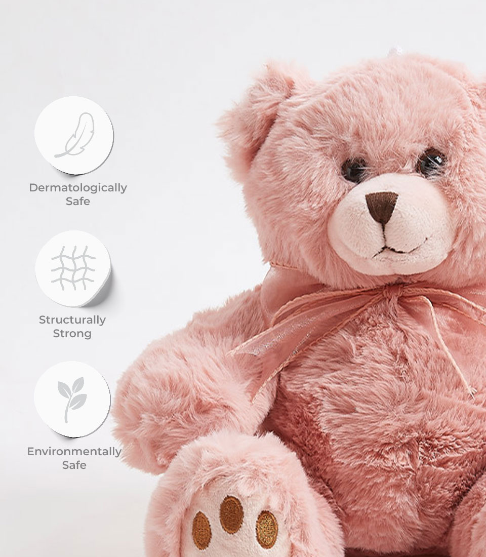 A plush Pink Teddy Bear with a bow around its neck sits against a plain background. Three icons near the bear indicate that it is dermatologically safe, structurally strong, and environmentally safe. The icons feature a feather, a network grid, and a leaf, respectively.