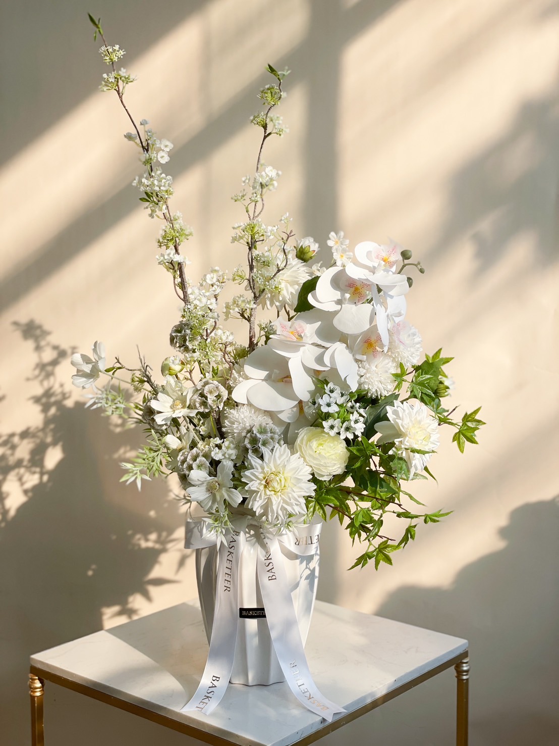 A sophisticated white floral arrangement featuring orchids, cherry blossoms, and lush greenery in a designer vase.