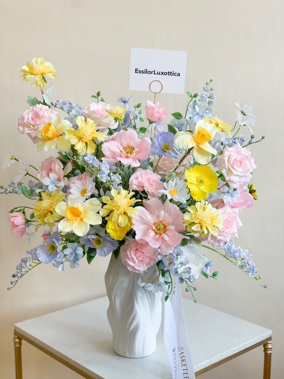 A large, vibrant floral arrangement sits in a white ceramic vase on a small table. This energetic spring bloom features pink roses, yellow and white daisies, blue hydrangeas, and purple delphiniums. A small card reading 