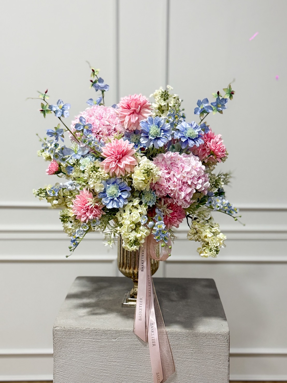 A floral arrangement in a gold Emerald Morning Renewal Artificial Flower Vase featuring pink, blue, and white flowers, including hydrangeas and dahlias, with greenery and a pink ribbon, displayed on a grey pedestal against a white panelled wall.