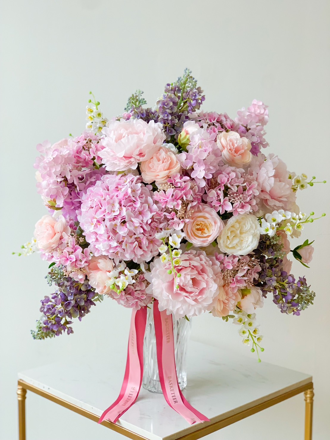 A large, lush bouquet of mixed flowers, including pink peonies, roses, hydrangeas, and purple lilacs, arranged in a clear glass vase. The vase sits on a white marble surface with gold trim. Two pink ribbons with white text hang from the bouquet, embodying graceful luxury.