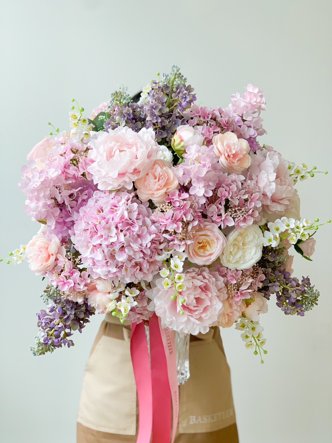 A graceful luxury bouquet of flowers arranged in soft pastel shades, including pink, white, and lavender blooms. The bouquet comprises roses, hydrangeas, and other mixed flowers adorned with green foliage. This elegant arrangement is tied with a pink ribbon and presented in an exquisite vase.