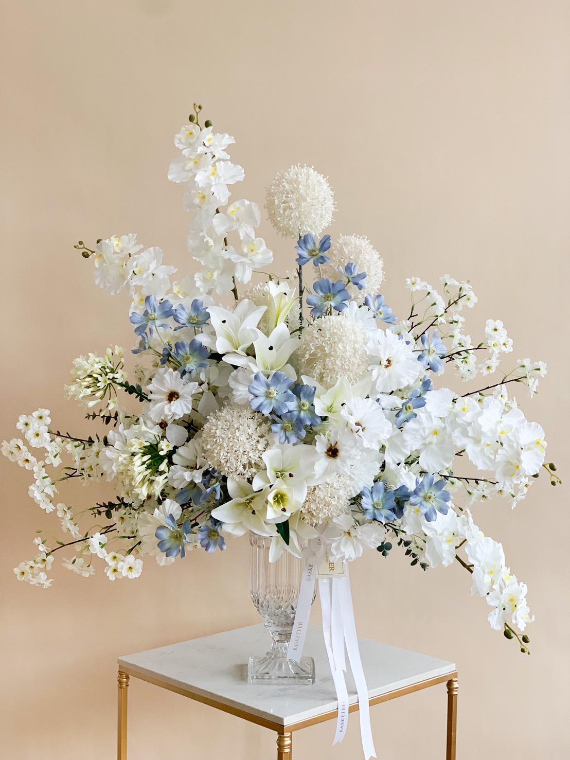 A large, elegant floral arrangement in a crystal vase. The bouquet features white lilies, hydrangeas, and delphiniums, accented with light blue flowers. White ribbons cascade from the ethereal winter floral display on a small white and gold table against a beige background.