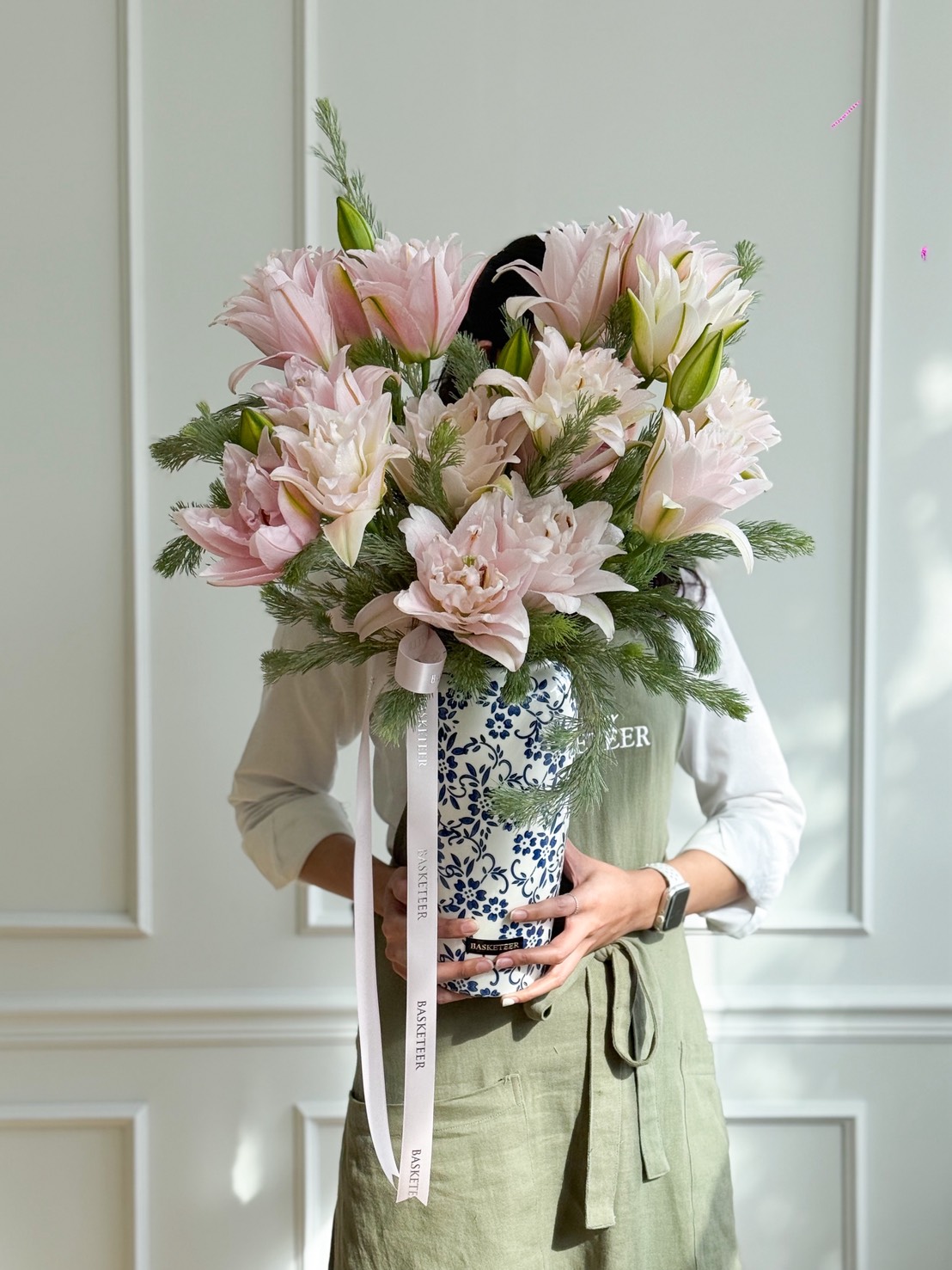A person in a green apron holds a large blue-and-white Artificial Flower Vase filled with pale pink lilies and greenery. The Charming Lily Elegance bouquet is adorned with a white ribbon featuring text, set against a white panelled wall.